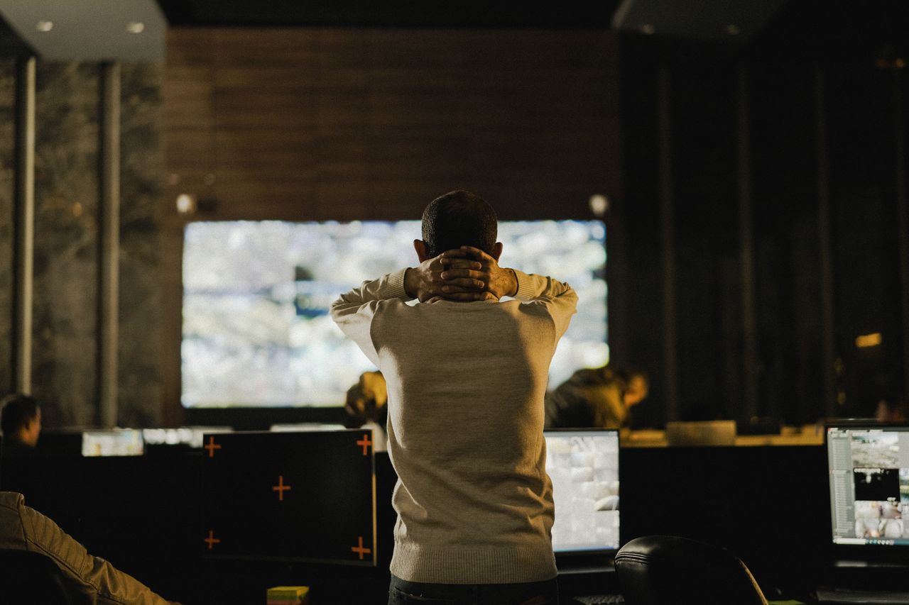 our-experience A man stands thoughtfully in a dimly lit control room with various screens.